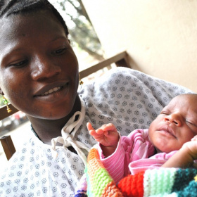 A picture of a young girl in a hospital gown holding a new born baby wrapped in a blanket and looking down at the child.