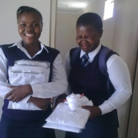 Two girls in school uniform standing next to each other and holding new white shirts in their hands. The girl on the left is smiling widely and the girl on the right is laughing with her eyes shut.
