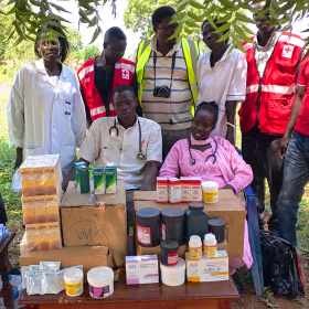 Five people wearing medical clothes are standing in a row, with two people wearing stethoscopes around their necks sat on chairs in front of them. In front of the whole group is a table piled high with boxes ad tubs of medicines.