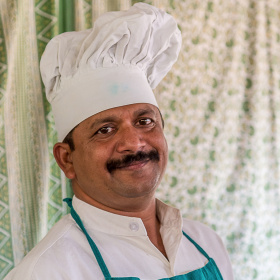 A headshot of a man with a moustache wearing a chef's hat. He is smiling and looks very happy.