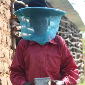 A picture of an elderly man wearing a big, black hat with a net hanging down covering his face. He is looking at the camera and holding a square object in his hands.