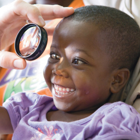 A close up picture of a very young child who is smiling widely. There is a hand holding an eye glass in front of one of her eyes.