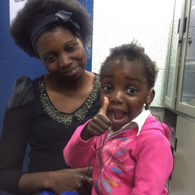 A mother is holding her young daughter who has hearing aids in her ears and is giving a big, excited thumbs up.