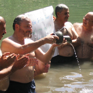 A picture of several men (mayors from Jordan, Israel and the Palestinian Authority) bathing in the Jordan river. They are holding up a sign, with some writing in English and some other scripts, and one of the men is holding a small metal jug and pouring w
