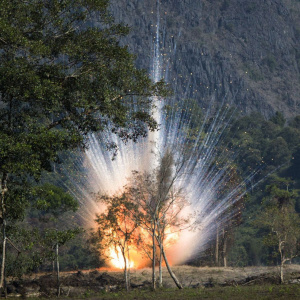 A photo of a large explosion, with sparks flying. In the background is a rock cliff face, and in the foreground some small trees.