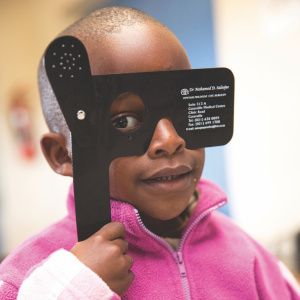 A close up of a young girl in a pink jumper, holding up a black glasses occluder to cover one of her eyes.