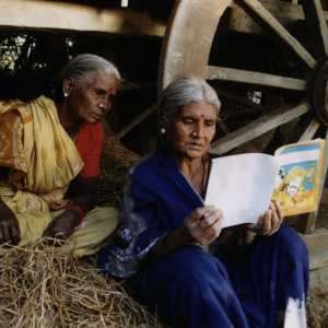 A picture of two elderly women sitting on a bale of hay looking at a book which one of the women is holding open.