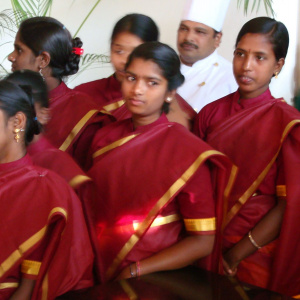 A photo of several young women in red and gold saris standing in a group, behind them is a man in chef's clothes.