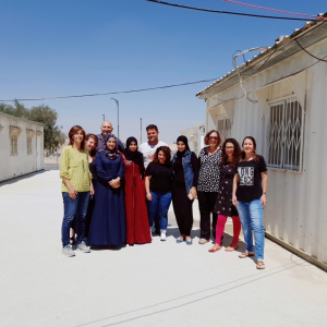 A picture of a group of people standing together in a bare street, with simple houses on each side. Some of them are wearing long dresses and hijabs and some of them are wearing jeans and t-shirts.
