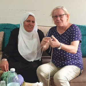 A picture of two women sitting on a sofa together, one wearing a hijab and the other dressed in Western clothes and holding a pair of knitting needles in her hands. 