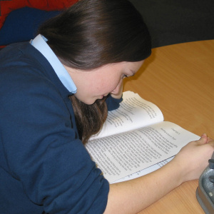 A picture of a teenage girl sat at a table bent over a book.