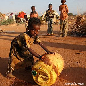 A picture of a young child in tattered clothes rolling a big plastic jug and looking at the camera. In the background there are three other children looking on and behind them what looks like a refugee camp.