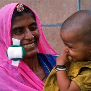 A close up picture of a mother holding a young child in her arms. Stuck to her cheek is a recently removed eye patch and she is smiling with joy.