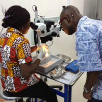 Two people are leaning over a small table with medical tools on it. The one on the left is looking through a microscope practising something on a plastic face mould, while the other is observing her.
