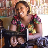 A picture of a woman at a table with a sewing machine on it. She is looking at the camera and smiling.