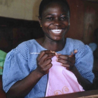 A picture of a young woman holding a pink cloth in her hands and a needle. She is smiling and looks very happy.