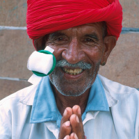 A close up picture of an older man with a eye patch stuck to his face, which has been removed to show his eye. He is smiling and holding his hand together in front of him.