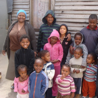 A group of children of varying ages are standing together in front of a wooden wall, with a woman next to them, who is smiling.