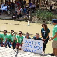 A group of young adults are kneeling in front of a murky looking river wearing matching t-shirts. Two of them are holding a sign saying 'Water cannot wait'. In the background, on the other side of the river are more people watching, one of them holding a 