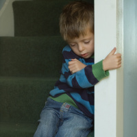 A picture of a young boy sat on a flight of stairs, holding onto the wall next to him tightly and looking sad.