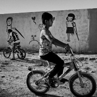 A black and white picture of a young boy on a bike, behind him is a wall with cartoon drawings of people who are missing limbs and using crutches.