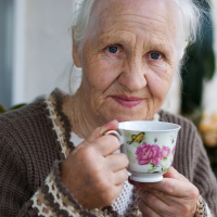 A close up picture of an elderly lady with grey hair holding a teacup up to her mouth and looking into the camera.