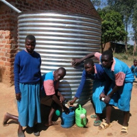 Four teenagers are standing in front of a large silver water tank, three of them are bent down filling up watering cans with water.