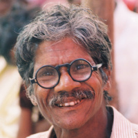 A headshot of an elderly man with greying hair and a moustache, wearing black-rimmed glasses and smiling.