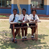Three teenage girls in school uniform sat at a desk outside, with notebooks on the table.